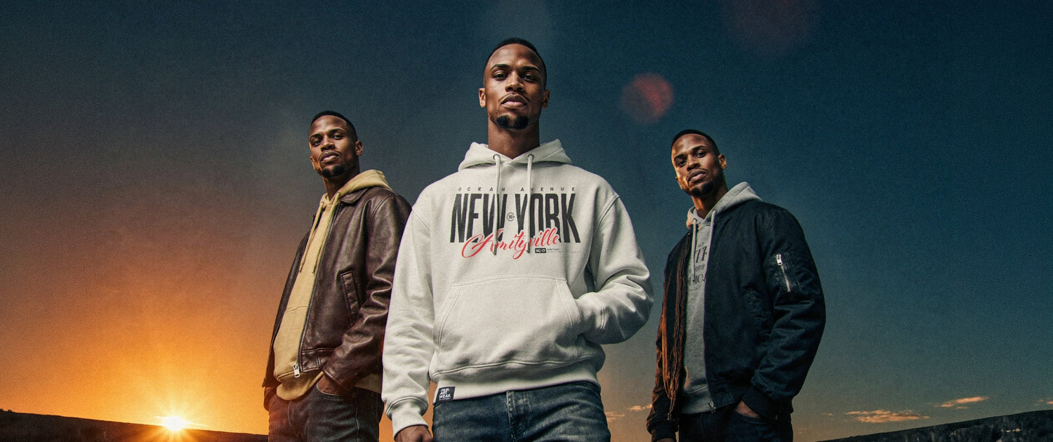 Organic Hoodie 'AMITYVILLE NY' – Drei Männer im cinematischen Sonnenuntergang-Setting, zentrale Figur trägt grauen Oversize Hoodie mit großflächigem Frontprint 'New York Amityville'. Dramatische Stimmung, filmisches Licht. Premium Cinematic Streetwear. Film Art Wear – Designed in Hamburg.
