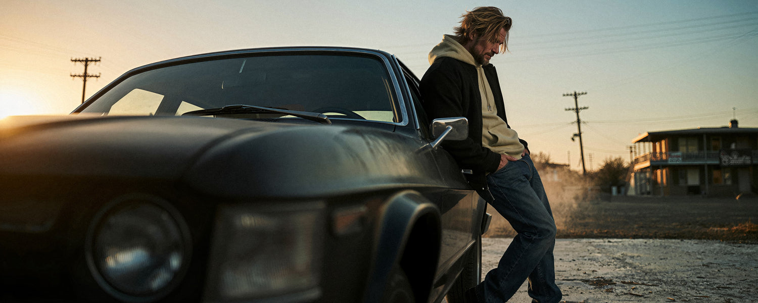 Mann lehnt an einem schwarzen Muscle Car bei Sonnenuntergang in verlassener Landschaft, trägt schwarzen Oversize Hoodie. Cinematische Weite, filmisches Roadmovie-Feeling. Premium Cinematic Streetwear. Film Art Wear – Designed in Hamburg.