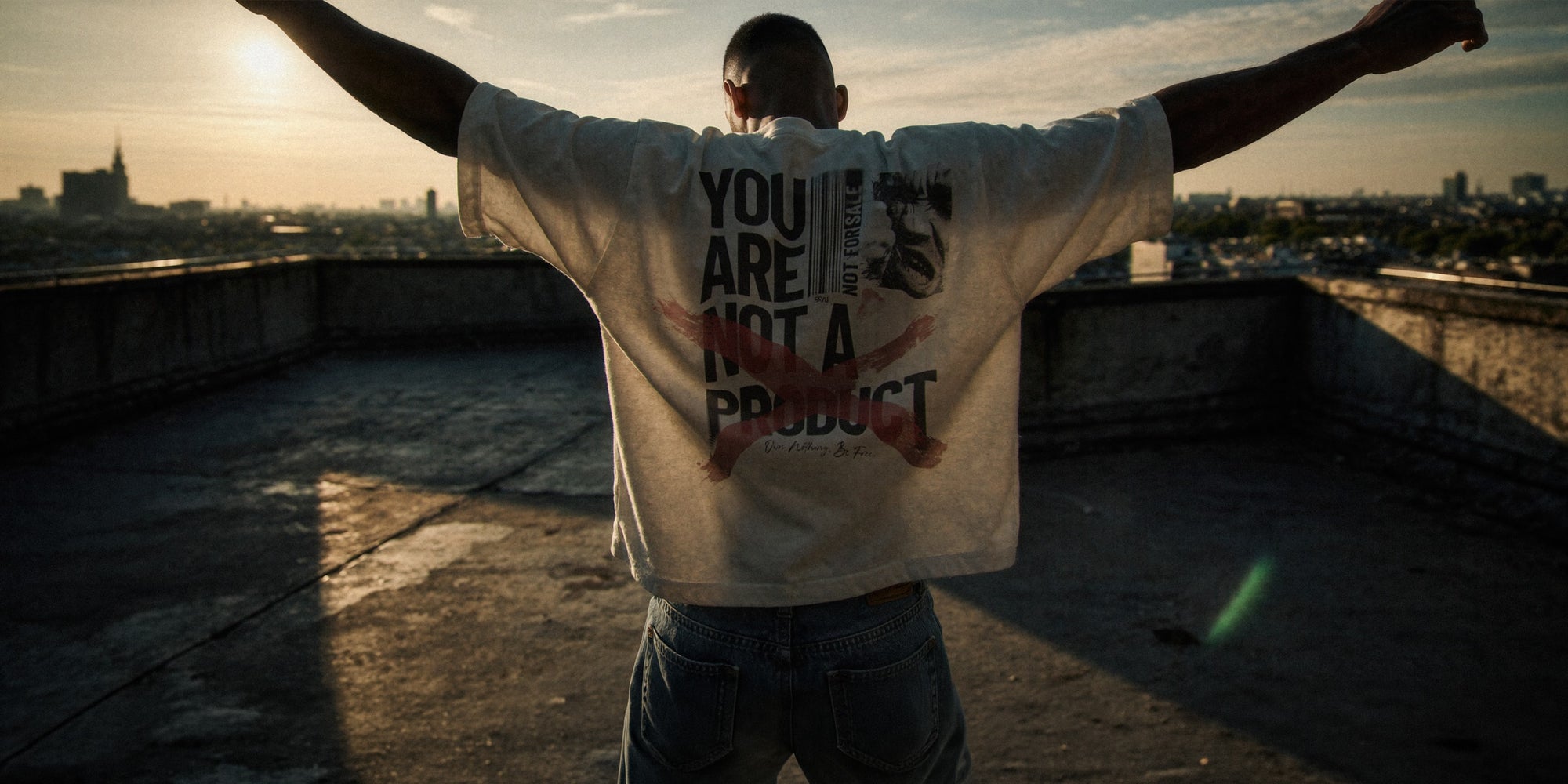 Mann steht mit ausgebreiteten Armen auf einem Rooftop mit Stadtpanorama bei Sonnenuntergang, trägt weißes Oversize T-Shirt 'NOT A PRODUCT' mit großflächigem Backprint 'You Are Not A Product'. Cinematische Freiheitsgeste. Premium Cinematic Streetwear. Film Art Wear – Designed in Hamburg.
