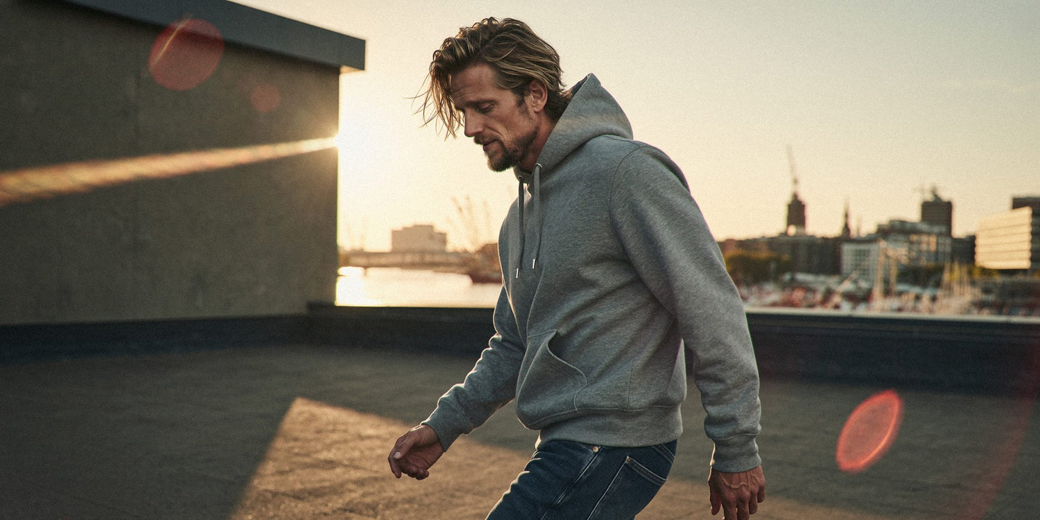 Mann geht über einen Rooftop mit Hamburger Hafenpanorama bei Sonnenuntergang, trägt grauen Oversize Hoodie. Cinematisches Gegenlicht, goldene Stunde. Premium Cinematic Streetwear. Film Art Wear – Designed in Hamburg.