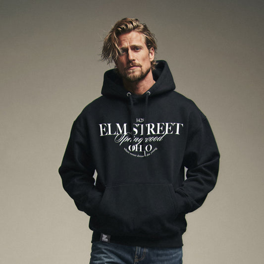 Organic Hoodie 'ELM STREET' – Mann von vorne, schwarzer Hoodie mit Frontprint '1428 Elm Street Springwood Ohio'. Iconic Places Design. Premium Cinematic Streetwear. Film Art Wear – Designed in Hamburg.