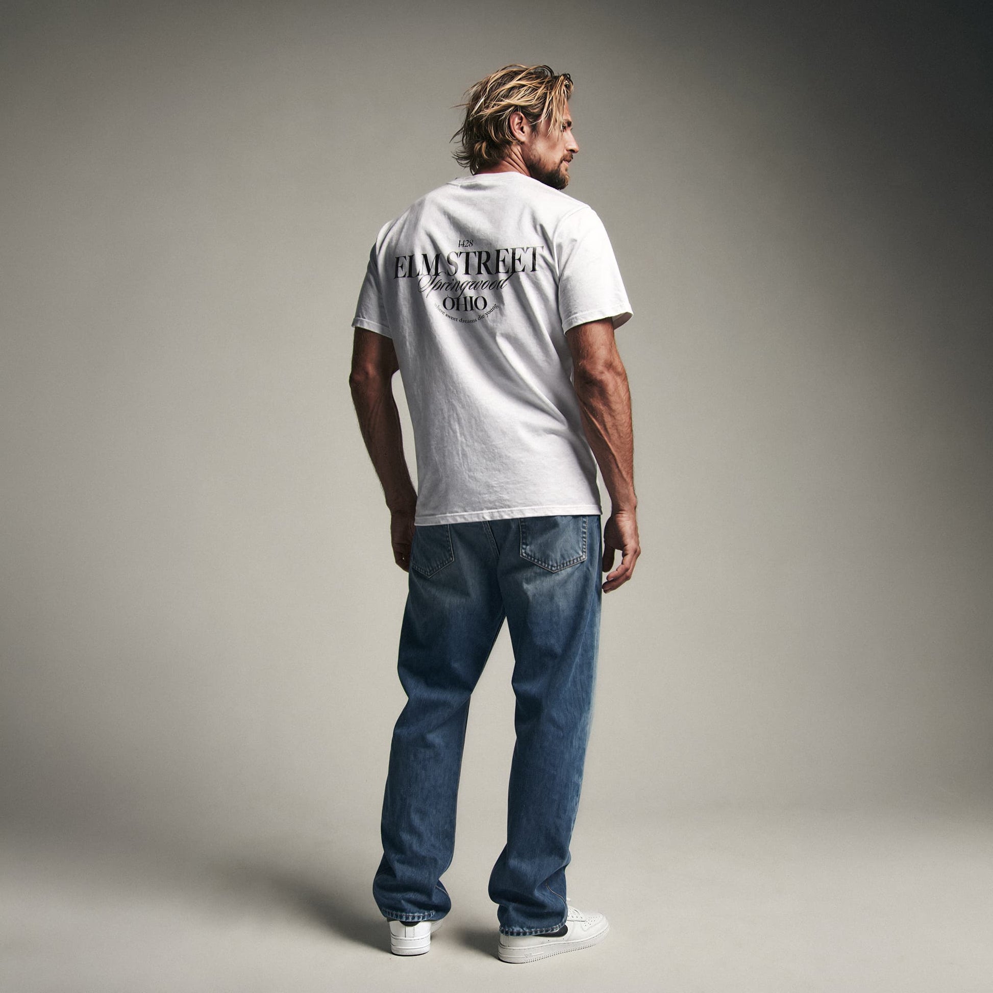 Organic T-Shirt 'ELM STREET' – Mann von hinten, weißes T-Shirt mit Backprint '1428 Elm Street Springwood Ohio'. Iconic Places Design. Premium Cinematic Streetwear. Film Art Wear – Designed in Hamburg.