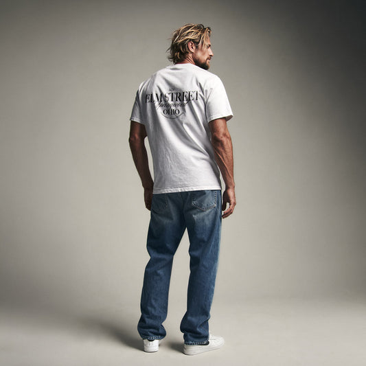 Organic T-Shirt 'ELM STREET' – Mann von hinten, weißes T-Shirt mit Backprint '1428 Elm Street Springwood Ohio'. Iconic Places Design. Premium Cinematic Streetwear. Film Art Wear – Designed in Hamburg.