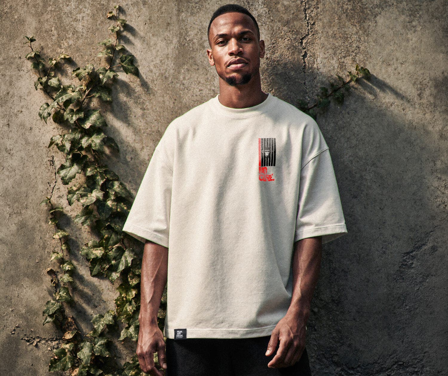 Mann steht aufrecht vor einer rohen Betonwand, trägt cremefarbenes Oversize T-Shirt aus 100% Bio-Baumwolle. Direkter Blick in die Kamera, Efeu-Ranke links im Bild. Urban, rau, ohne Kompromisse. Premium Cinematic Streetwear. Film Art Wear – Designed in Hamburg.