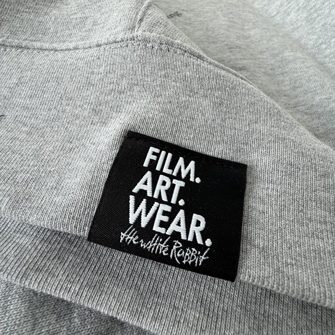 Extreme Nahaufnahme des eingenähten Film Art Wear Labels am Saum eines grauen Hoodies. Schwarzes Webetikelt mit weißer Stickerei 'Film. Art. Wear.' Qualitätsdetail. Film Art Wear – Designed in Hamburg.